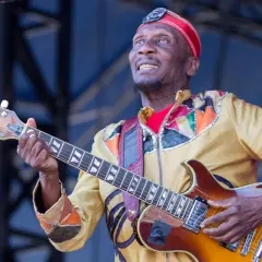 Ein verstorbener Musiker, eine Legende, spielt Gitarre auf der Bühne. Sein buntes Outfit und sein Ausdruck zeugen von seiner Leidenschaft.