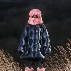 Ein Model in einem voluminösen, schwarzen Daunenmantel mit pinkem Schal und Mütze, inspiriert von Dingyun Zhang, steht in trockenem Gras.