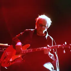 Jack Casady, mit seinem charakteristischen weißen Haar und Sonnenbrille, spielt auf einer roten E-Gitarre, während rotes Licht die Bühne erhellt.