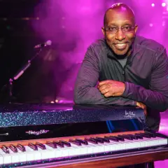 Greg Phillinganes lächelt, lehnt sich an ein glitzerndes Klavier auf einer Bühne im lila Licht.