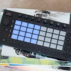 Ein schwarzes Ableton-Controller-Gerät mit leuchtenden blauen Pads liegt auf gestapelten Büchern.