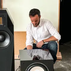 Mann im weißen Hemd und Jeans macht einen **subwoofer test** vor einem großen schwarzen Lautsprecher.