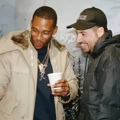 Ronnie Fieg und ein Freund schauen auf ein Handy, während sie lächelnd und in Winterjacken vor einer Bergkulisse stehen.