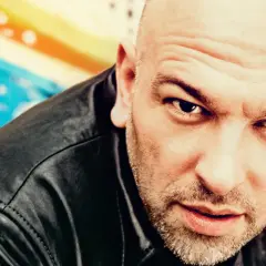 Pascal Feos, ein Mann mit Glatze und Bart, blickt intensiv in die Kamera. Sein schwarzes Lederoutfit und der bunte Hintergrund erzeugen einen künstlerischen Eindruck.