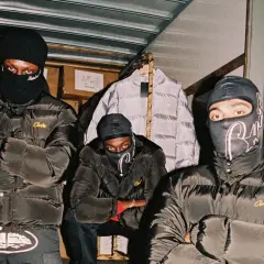 Drei Männer in schwarzen Daunenjacken mit dem **Corteiz logo** und Sturmhauben posieren vor einem Lagerhaus.