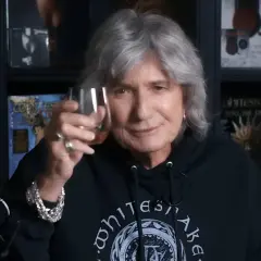 David Coverdale heute, mit grauem Haar und einem Glas in der Hand, trägt ein schwarzes Whitesnake-Hoodie.