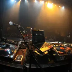 DJ mit roter Kapuze legt auf. Scheinwerfer beleuchten die Bühne und das Publikum. Die Namen der DJ-Größen sind auf den Platten zu sehen.