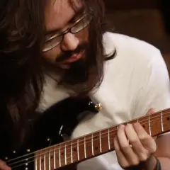 Mateus Asato, mit langen Haaren und Brille, spielt konzentriert auf seiner E-Gitarre.