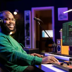 Ein Mann mit Kopfhörern spielt auf einem Keyboard in einem Musikstudio. Auf dem Monitor sind Wellenformen und Regler zu sehen, wie bei Konzeptkunst-Beispielen.