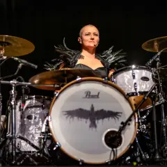 Katha Mia, die Schlagzeugerin, mit Federkragen und einem Raben-Logo auf der Bassdrum, bereit für ihren Auftritt.