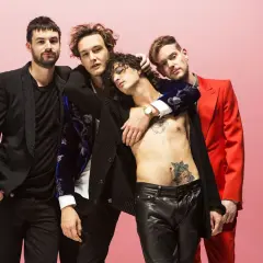 Die Mitglieder von The 1975 posieren eng umschlungen vor einem rosafarbenen Hintergrund. Einer trägt einen roten Anzug, ein anderer ein schwarzes Jackett.