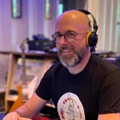Hans Martin Buff, mit Kopfhörern und Brille, lächelt in einem Tonstudio. Im Hintergrund sind Gitarren und Musik-Equipment zu sehen.