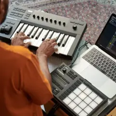 Mann spielt auf einem Keyboard, daneben ein Laptop mit Musiksoftware und ein Pad-Controller. Native Instruments-Geräte für kreative Musikproduktion.