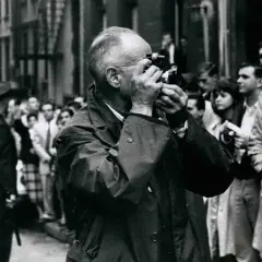 Ein Mann mit Brille fotografiert mit einer Leica, umgeben von einer Menge. Ein typisches Bild, das an Henri Cartier-Bresson Fotos erinnert.