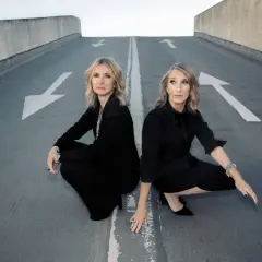 Susanne Freytag und ihre Partnerin, beide in eleganten schwarzen Outfits, hocken auf einer asphaltierten Rampe mit weißen Pfeilen.