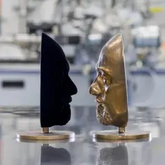Zwei Gesichtsmasken stehen sich gegenüber: eine aus samtigem, dunkelstem Schwarz, die andere aus glänzendem Bronze.