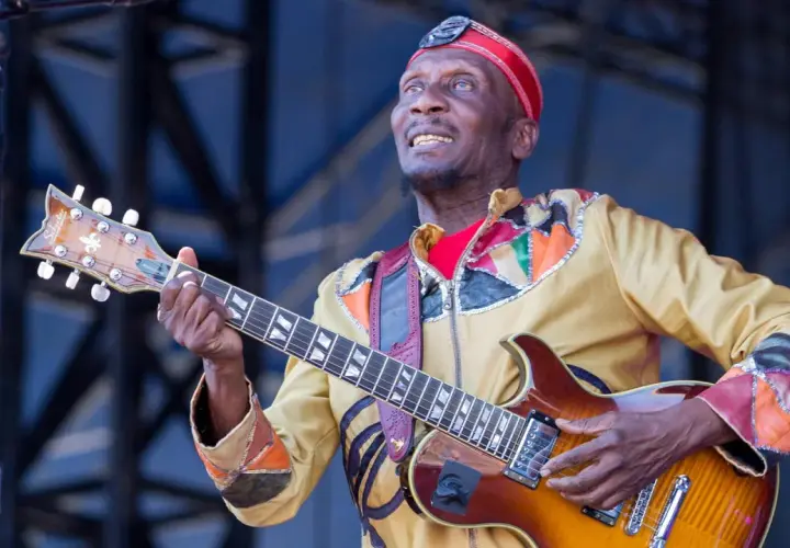 Ein verstorbener Musiker, eine Legende, spielt Gitarre auf der Bühne. Sein buntes Outfit und sein Ausdruck zeugen von seiner Leidenschaft.