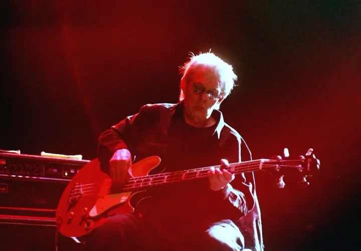 Jack Casady, mit seinem charakteristischen weißen Haar und Sonnenbrille, spielt auf einer roten E-Gitarre, während rotes Licht die Bühne erhellt.