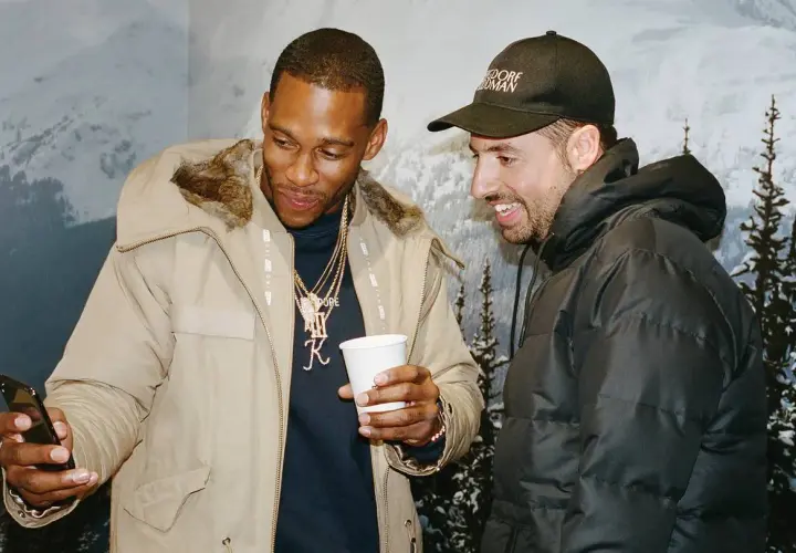 Ronnie Fieg und ein Freund schauen auf ein Handy, während sie lächelnd und in Winterjacken vor einer Bergkulisse stehen.