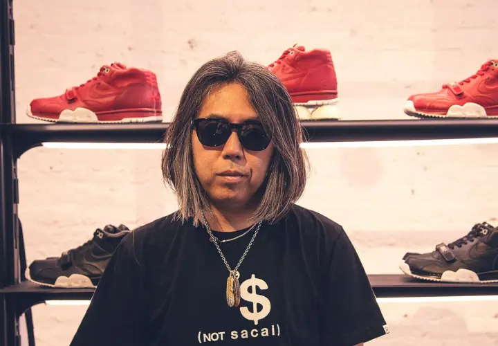 Mann mit Sonnenbrille und Kette vor roten und schwarzen Sneakern. Sein T-Shirt zeigt ein Dollarzeichen und den Schriftzug "(not sacai)", ein Hinweis auf die fragment brand.