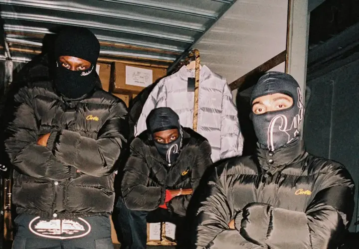 Drei Männer in schwarzen Daunenjacken mit dem **Corteiz logo** und Sturmhauben posieren vor einem Lagerhaus.