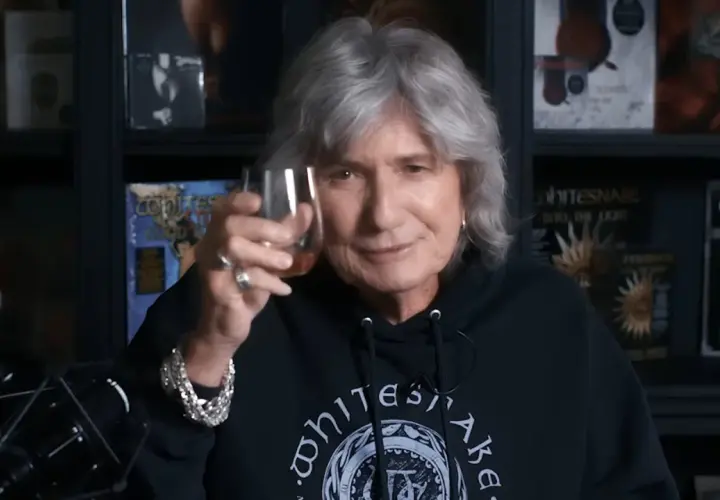 David Coverdale heute, mit grauem Haar und einem Glas in der Hand, trägt ein schwarzes Whitesnake-Hoodie.
