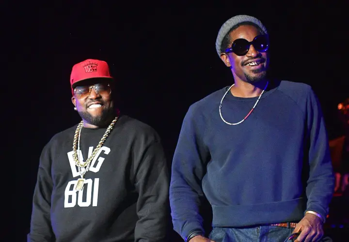 Die Mitglieder von Outkast, Big Boi mit roter Kappe und Andre 3000 mit grauer Mütze, lächeln auf der Bühne.