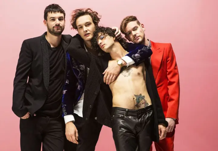 Die Mitglieder von The 1975 posieren eng umschlungen vor einem rosafarbenen Hintergrund. Einer trägt einen roten Anzug, ein anderer ein schwarzes Jackett.