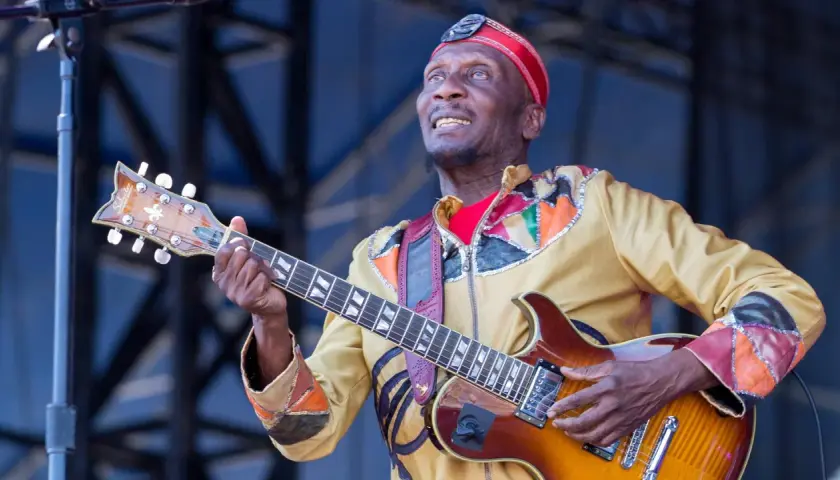 Ein verstorbener Musiker, eine Legende, spielt Gitarre auf der Bühne. Sein buntes Outfit und sein Ausdruck zeugen von seiner Leidenschaft.