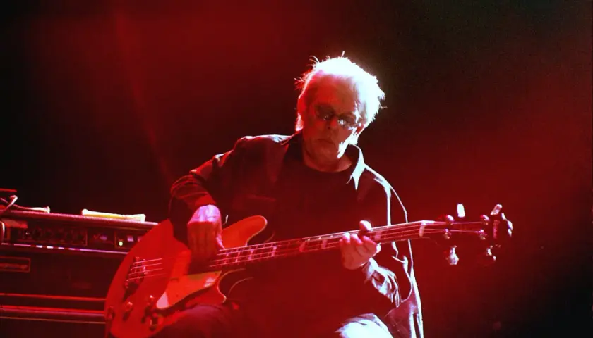 Jack Casady, mit seinem charakteristischen weißen Haar und Sonnenbrille, spielt auf einer roten E-Gitarre, während rotes Licht die Bühne erhellt.