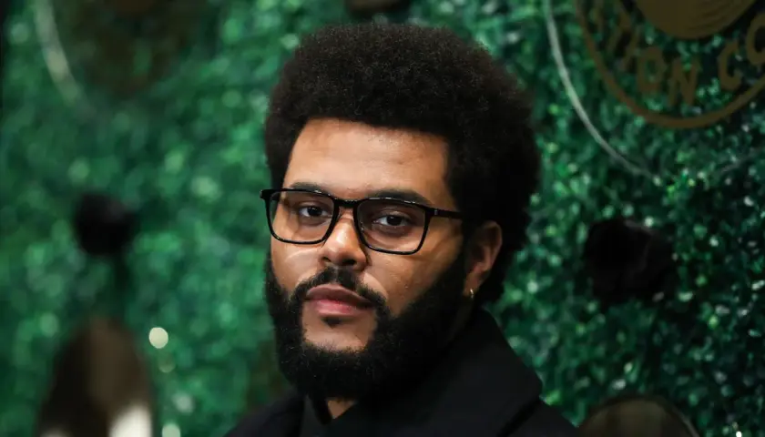 The Weeknd, mit Brille und Bart, posiert vor einer grünen Wand. Seine Musikkarriere begann in Toronto, Kanada.