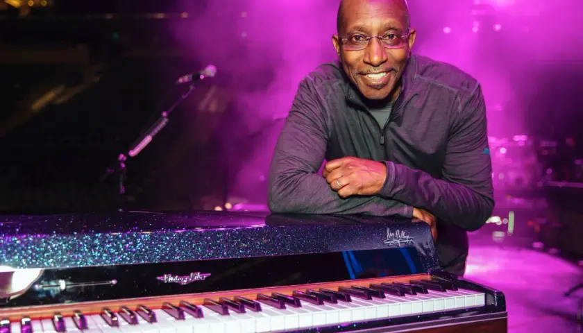 Greg Phillinganes lächelt, lehnt sich an ein glitzerndes Klavier auf einer Bühne im lila Licht.