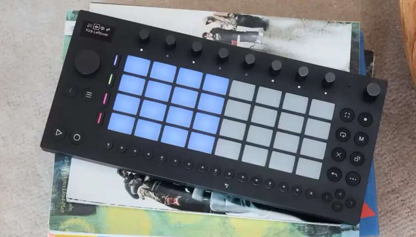 Ein schwarzes Ableton-Controller-Gerät mit leuchtenden blauen Pads liegt auf gestapelten Büchern.
