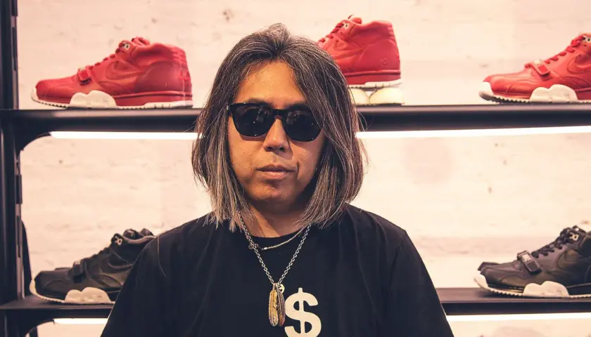 Mann mit Sonnenbrille und Kette vor roten und schwarzen Sneakern. Sein T-Shirt zeigt ein Dollarzeichen und den Schriftzug "(not sacai)", ein Hinweis auf die fragment brand.