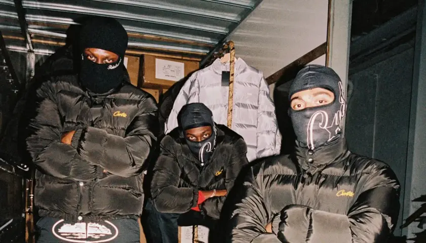 Drei Männer in schwarzen Daunenjacken mit dem **Corteiz logo** und Sturmhauben posieren vor einem Lagerhaus.