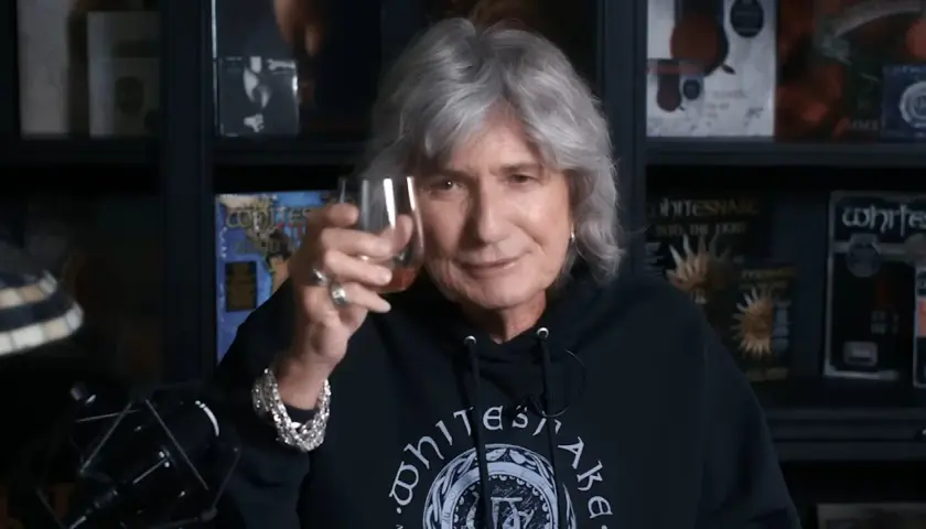David Coverdale heute, mit grauem Haar und einem Glas in der Hand, trägt ein schwarzes Whitesnake-Hoodie.