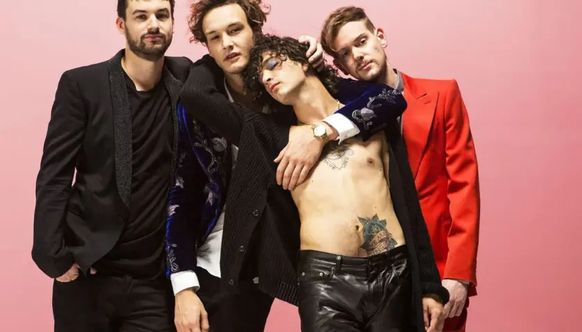 Die Mitglieder von The 1975 posieren eng umschlungen vor einem rosafarbenen Hintergrund. Einer trägt einen roten Anzug, ein anderer ein schwarzes Jackett.