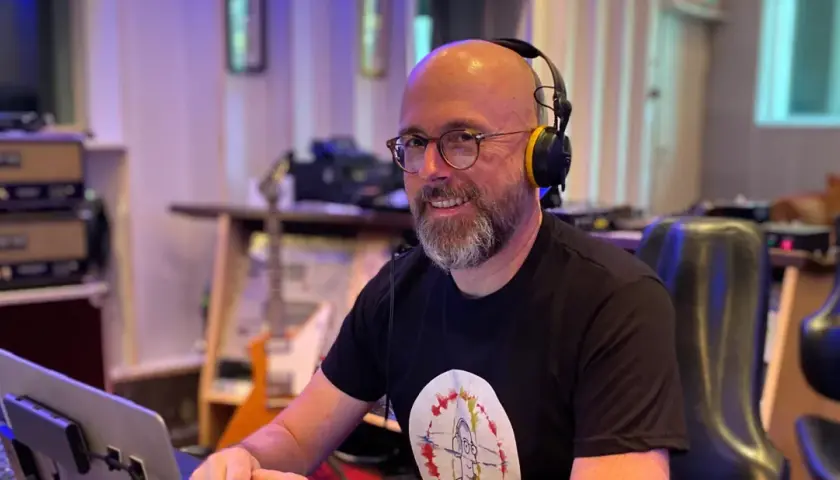 Hans Martin Buff, mit Kopfhörern und Brille, lächelt in einem Tonstudio. Im Hintergrund sind Gitarren und Musik-Equipment zu sehen.
