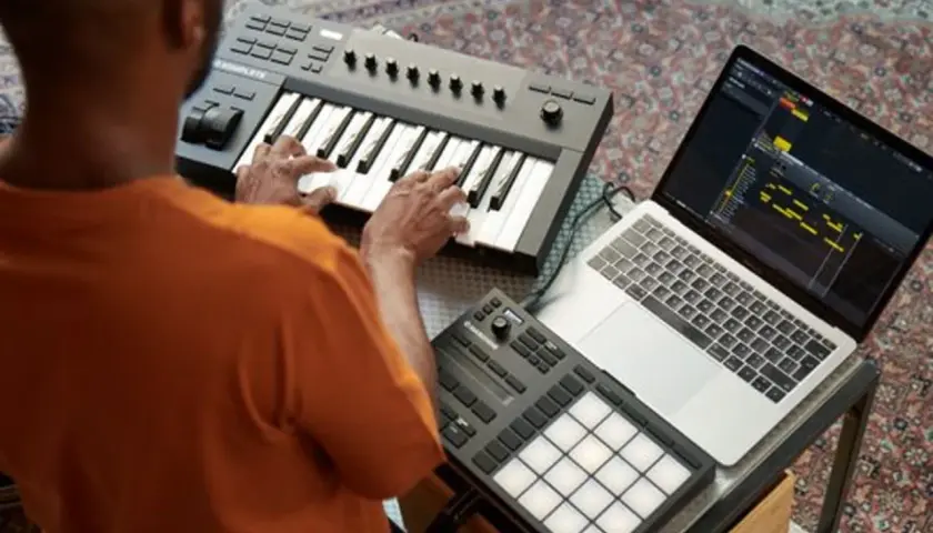 Mann spielt auf einem Keyboard, daneben ein Laptop mit Musiksoftware und ein Pad-Controller. Native Instruments-Geräte für kreative Musikproduktion.