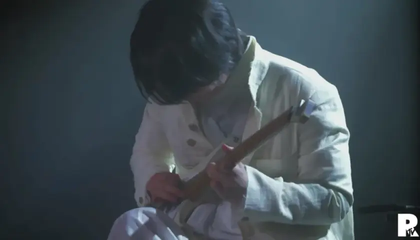 Ein Musiker in weißer Kleidung spielt eine Gitarre. Die Szene ist düster beleuchtet, was die Konzentration auf die Performance von Ichika Nito verstärkt.