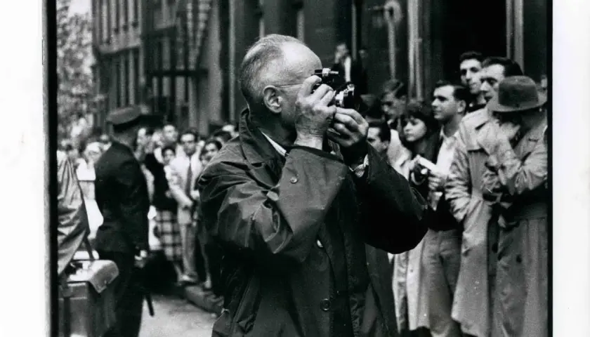 Ein Mann mit Brille fotografiert mit einer Leica, umgeben von einer Menge. Ein typisches Bild, das an Henri Cartier-Bresson Fotos erinnert.