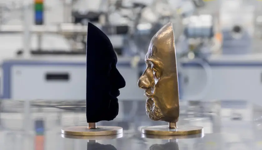 Zwei Gesichtsmasken stehen sich gegenüber: eine aus samtigem, dunkelstem Schwarz, die andere aus glänzendem Bronze.
