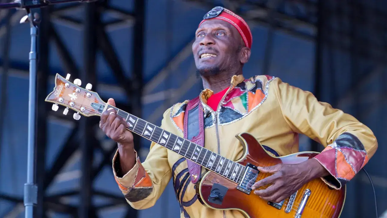 Ein verstorbener Musiker, eine Legende, spielt Gitarre auf der Bühne. Sein buntes Outfit und sein Ausdruck zeugen von seiner Leidenschaft.