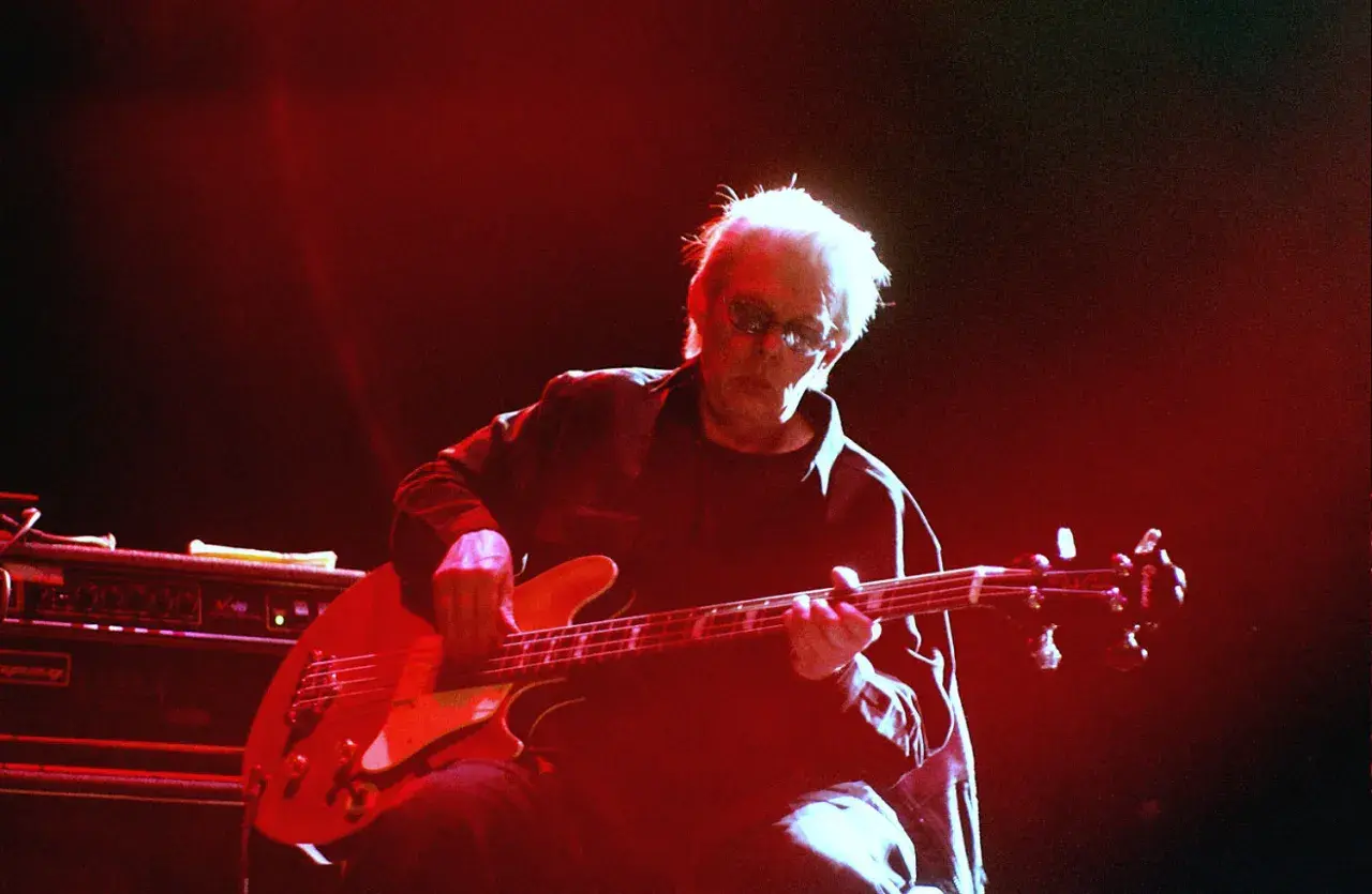Jack Casady, mit seinem charakteristischen weißen Haar und Sonnenbrille, spielt auf einer roten E-Gitarre, während rotes Licht die Bühne erhellt.