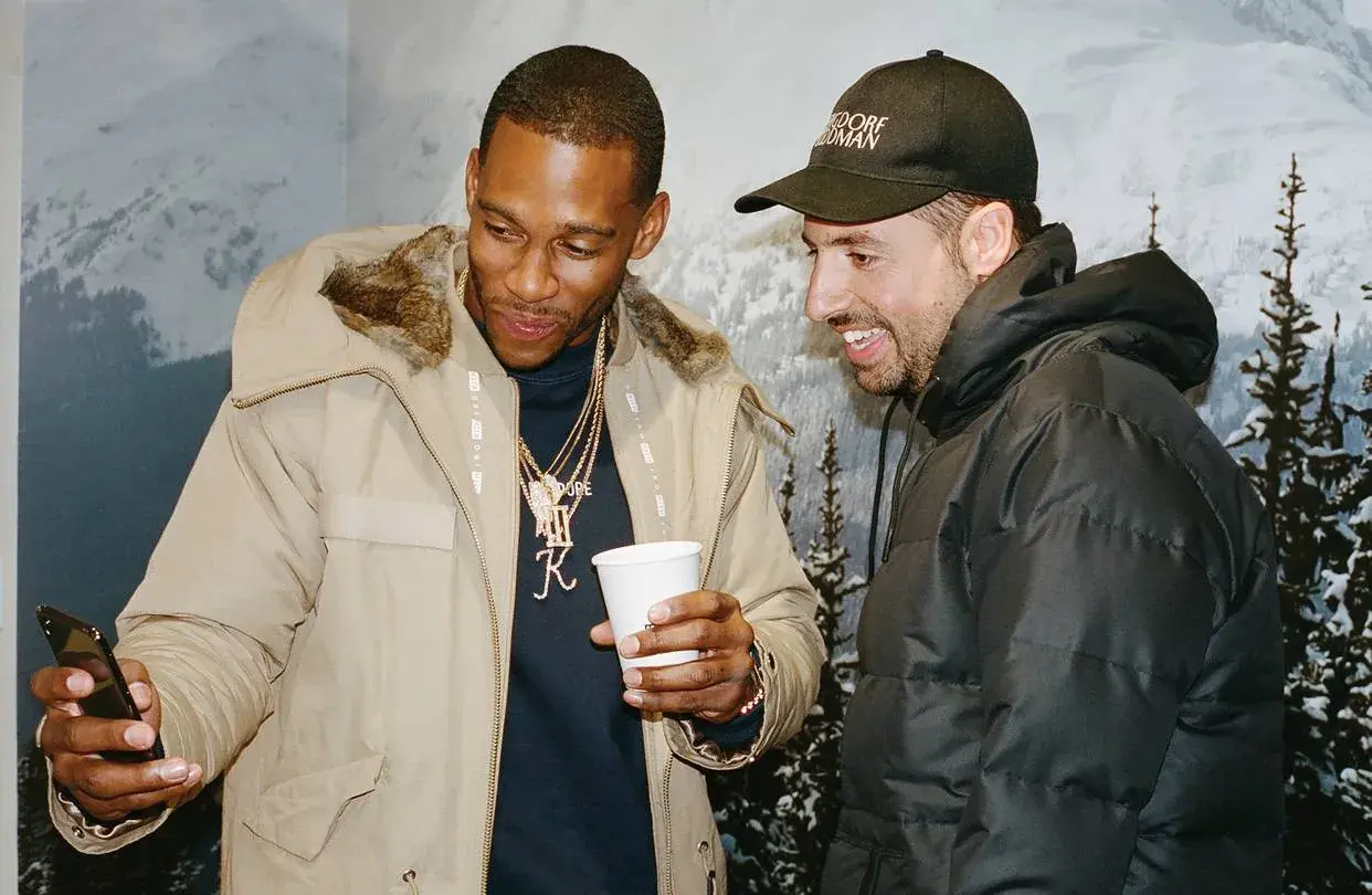 Ronnie Fieg und ein Freund schauen auf ein Handy, während sie lächelnd und in Winterjacken vor einer Bergkulisse stehen.