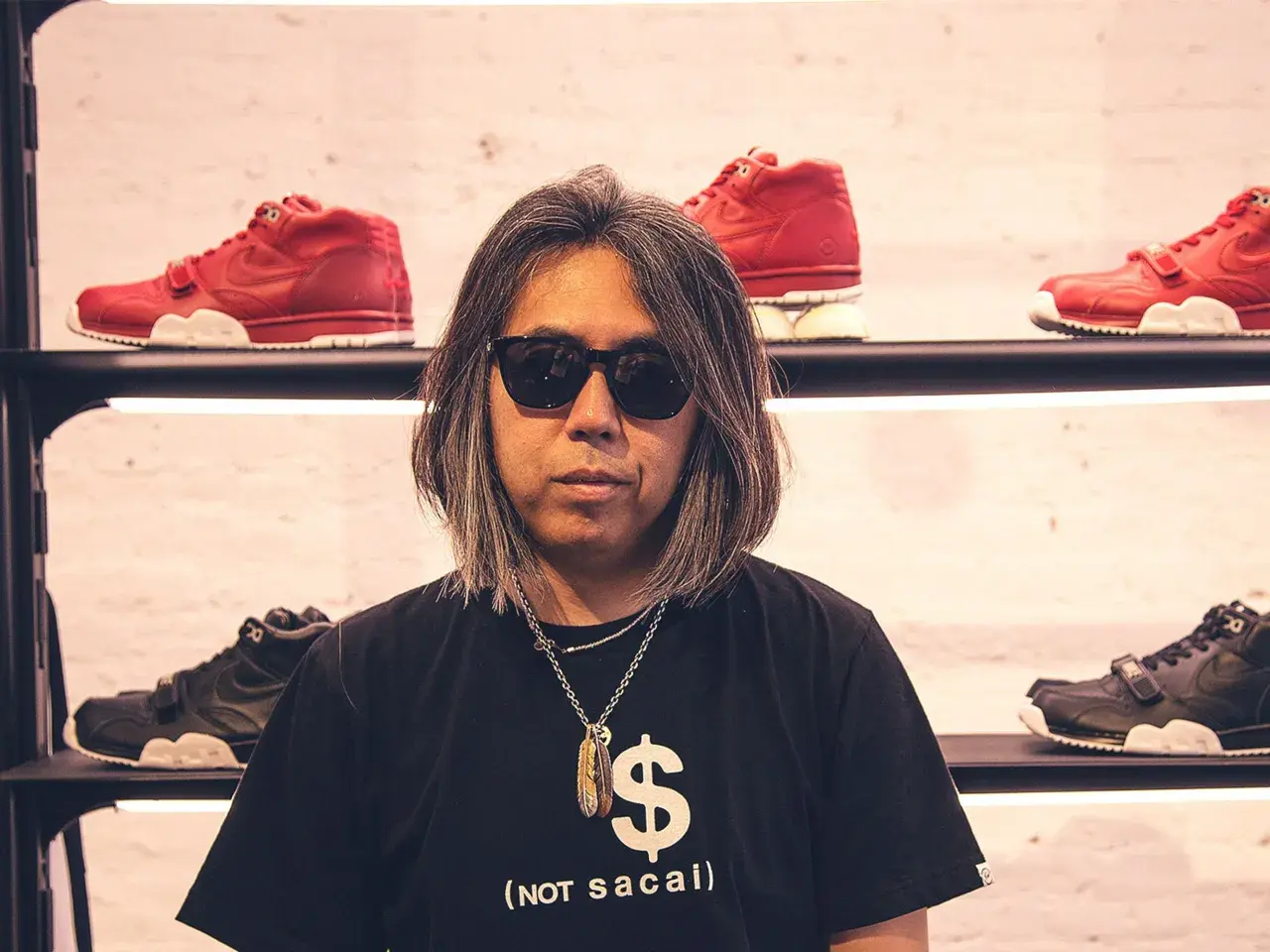 Mann mit Sonnenbrille und Kette vor roten und schwarzen Sneakern. Sein T-Shirt zeigt ein Dollarzeichen und den Schriftzug "(not sacai)", ein Hinweis auf die fragment brand.