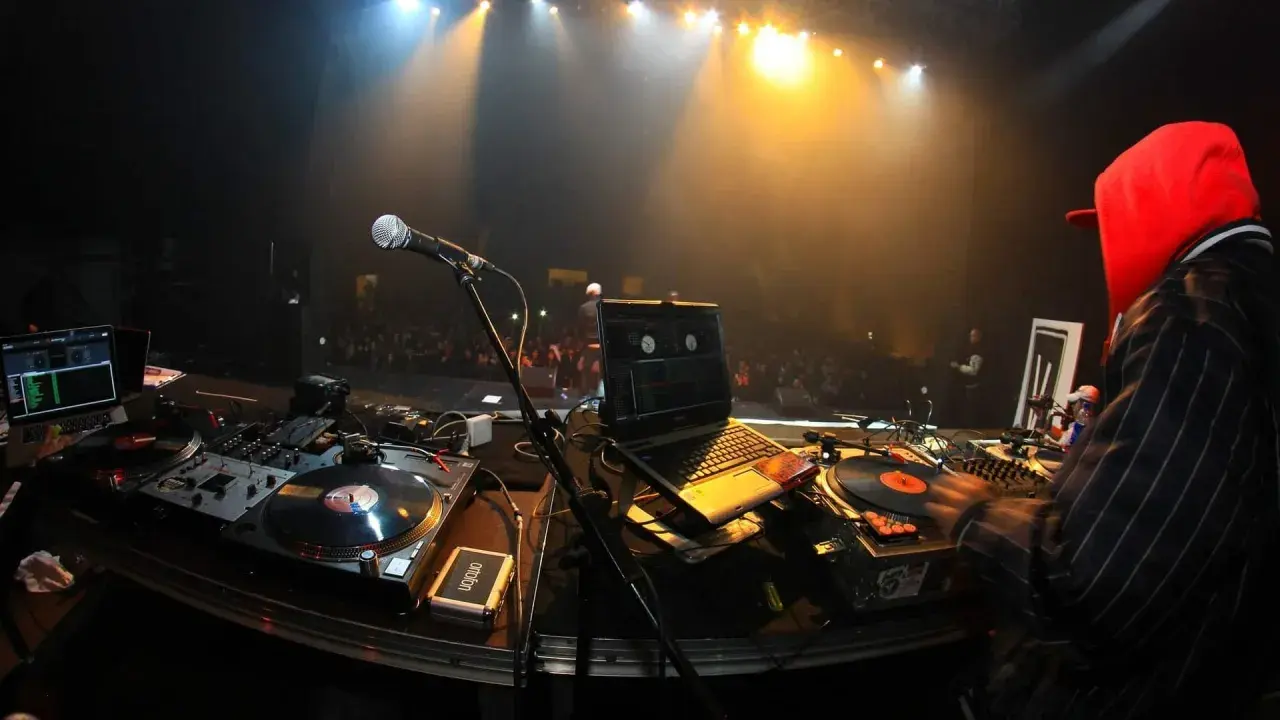 DJ mit roter Kapuze legt auf. Scheinwerfer beleuchten die Bühne und das Publikum. Die Namen der DJ-Größen sind auf den Platten zu sehen.