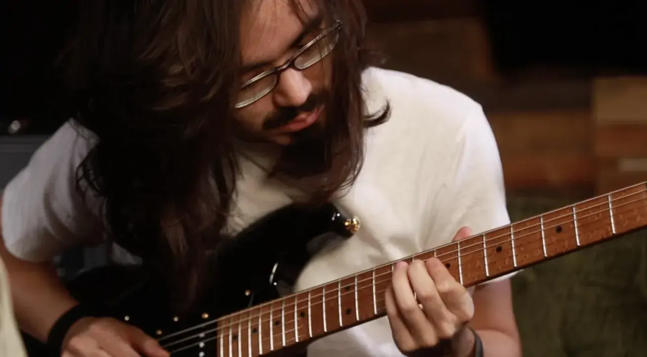 Mateus Asato, mit langen Haaren und Brille, spielt konzentriert auf seiner E-Gitarre.