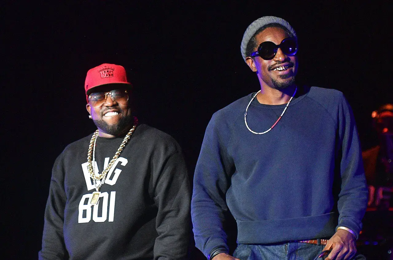 Die Mitglieder von Outkast, Big Boi mit roter Kappe und Andre 3000 mit grauer Mütze, lächeln auf der Bühne.