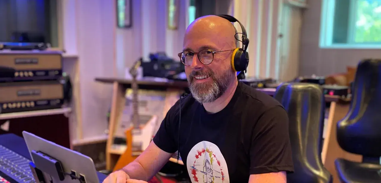 Hans Martin Buff, mit Kopfhörern und Brille, lächelt in einem Tonstudio. Im Hintergrund sind Gitarren und Musik-Equipment zu sehen.